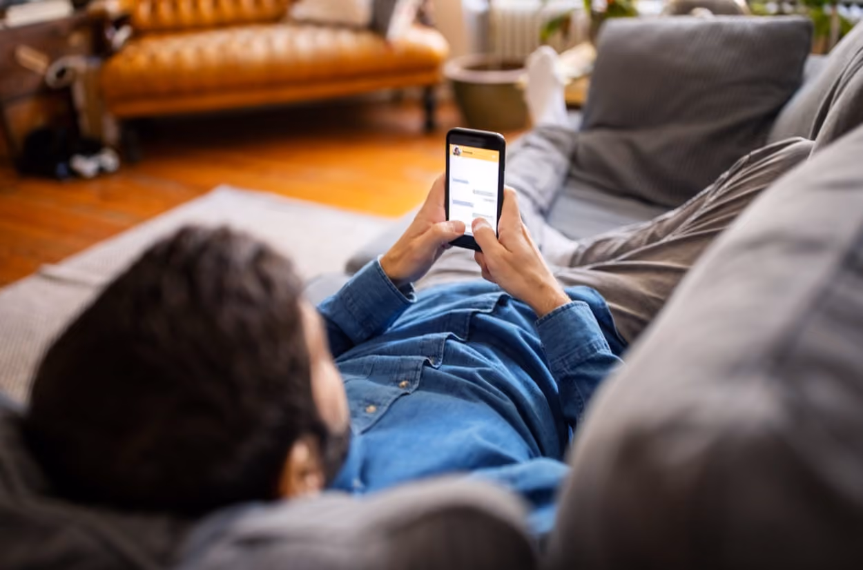 A man sitting on a sofa at home, focused on using his smartphone, surrounded by a cozy living room atmosphere.