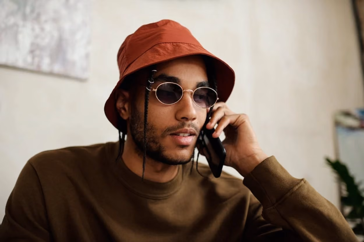 A man wearing a hat and sunglasses is engaged in a conversation on his cell phone.