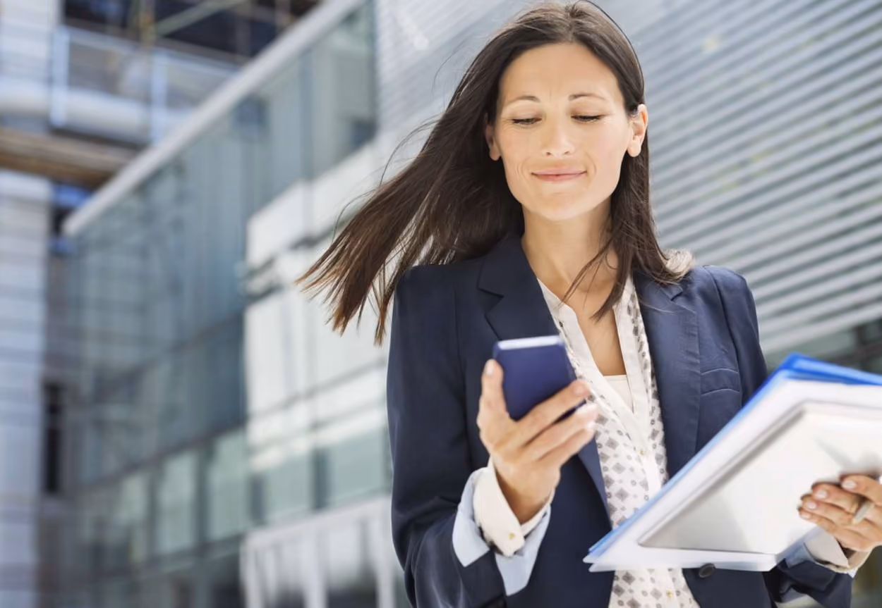 A businesswoman in an office setting, focused on her mobile phone while engaged in work-related tasks.