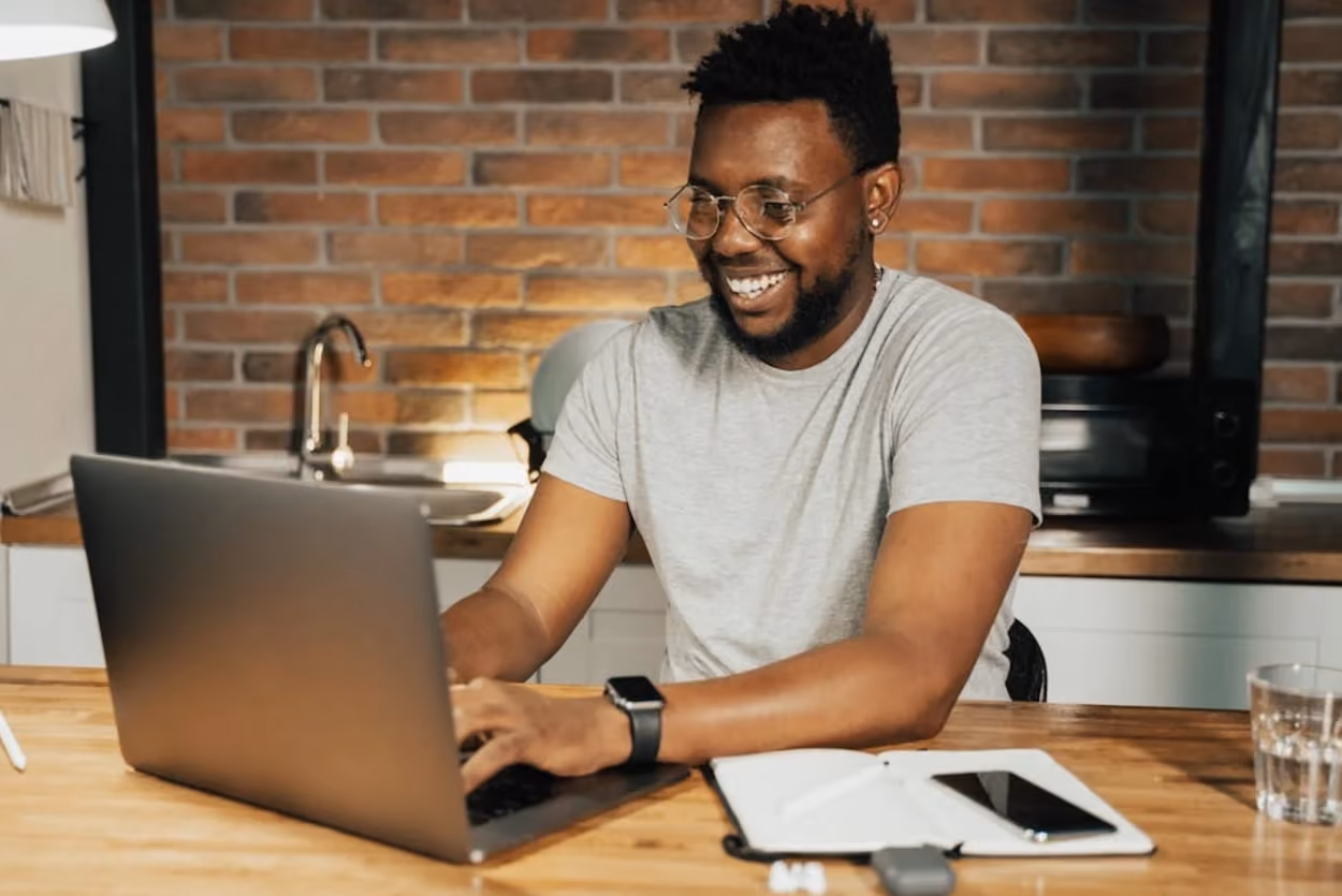 A smiling man sits at a table, engaged with his laptop, exuding a sense of contentment and focus.