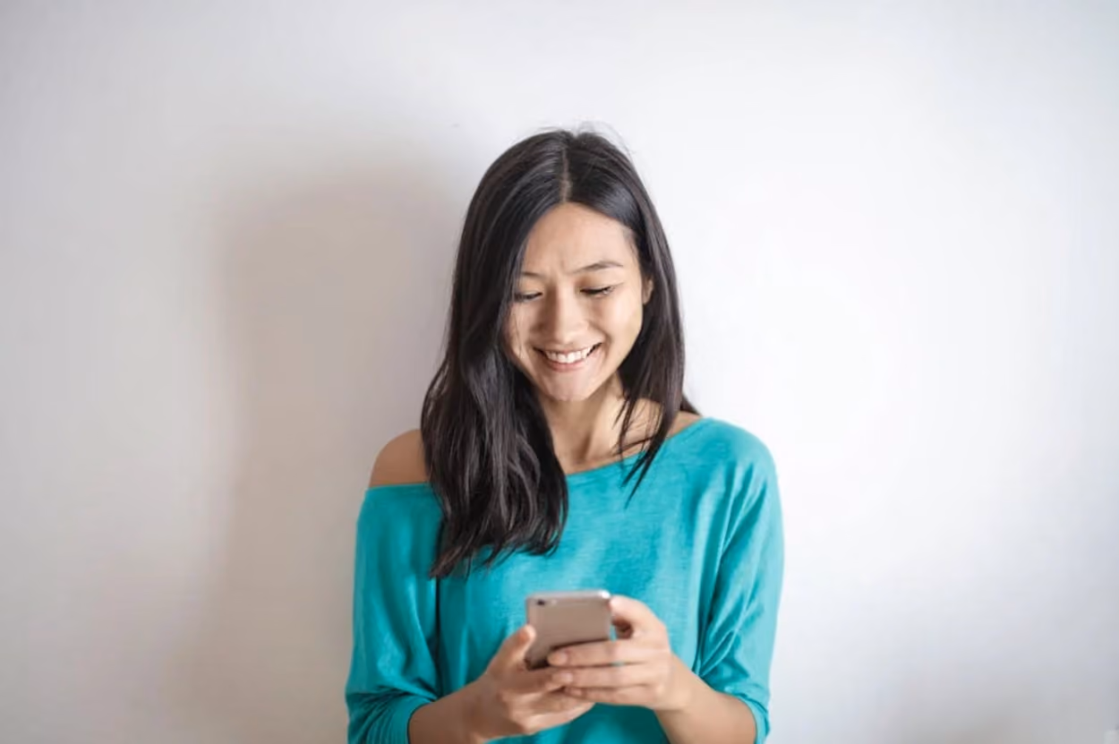 A woman smiles brightly while holding her phone, exuding joy and positivity in her expression.