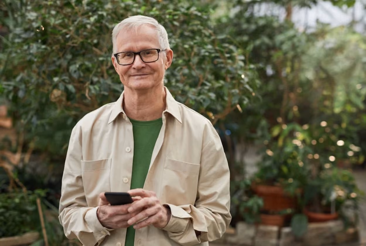 Senior man engaged with a smartphone while seated in a lush garden, surrounded by greenery and blooming flowers.