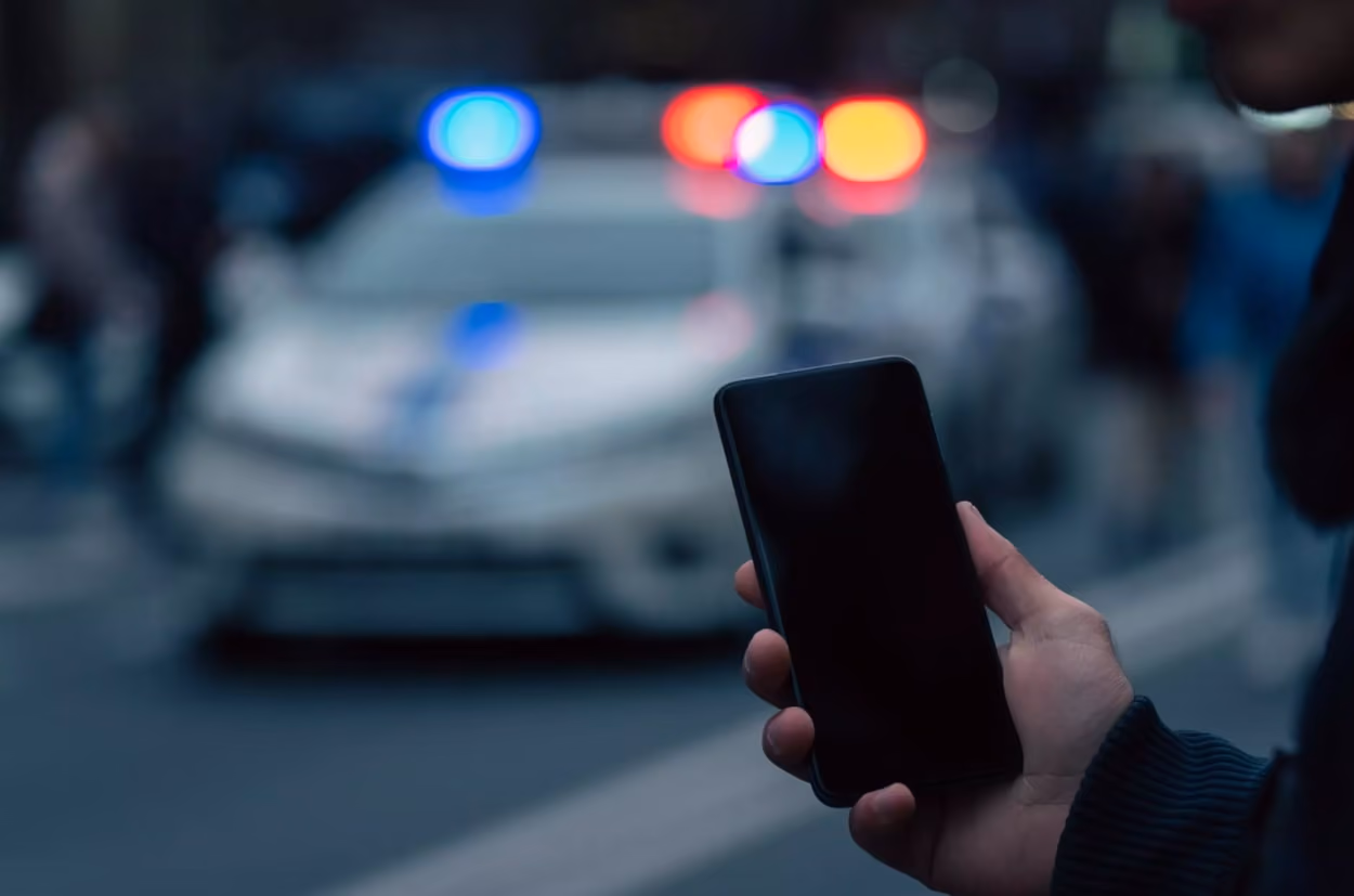 A person holding a cell phone, illuminated by flashing police lights in the background, conveying urgency and attention.