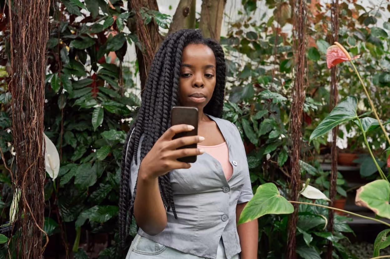 A woman stands in a garden, intently looking at her phone amidst blooming flowers and greenery.