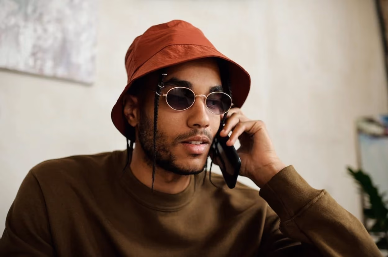 A man wearing a hat and sunglasses is engaged in a conversation on his cell phone.