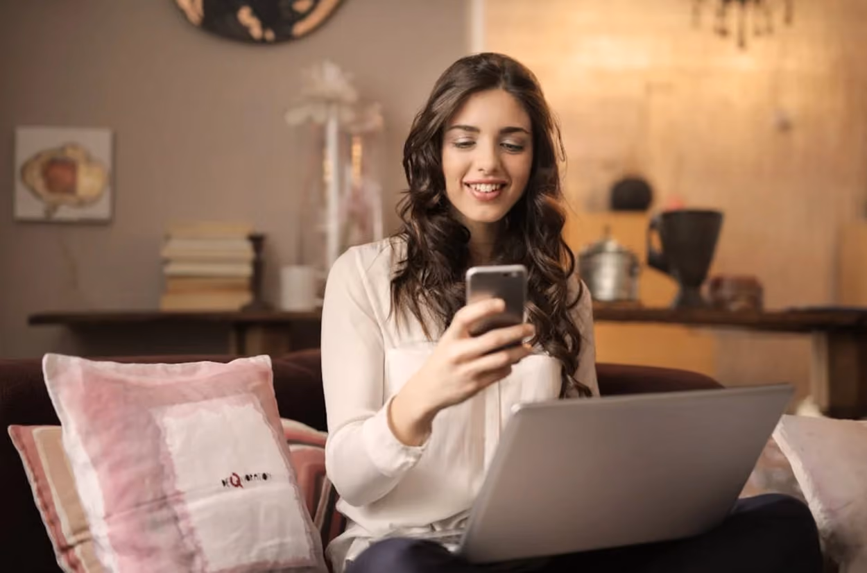 A woman seated on a couch, using a laptop while holding a cell phone in her hand, engaged in work or communication.