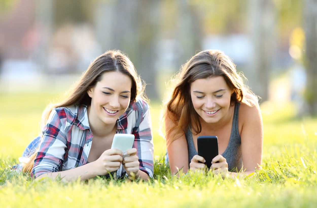 Two young women relax on the grass, each engaged with their cell phones, enjoying a sunny day outdoors.