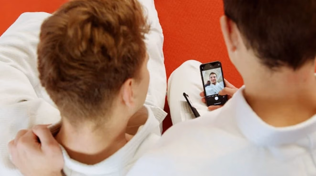 Two men in white robes sit on the floor; one is using a cell phone while the other looks on.