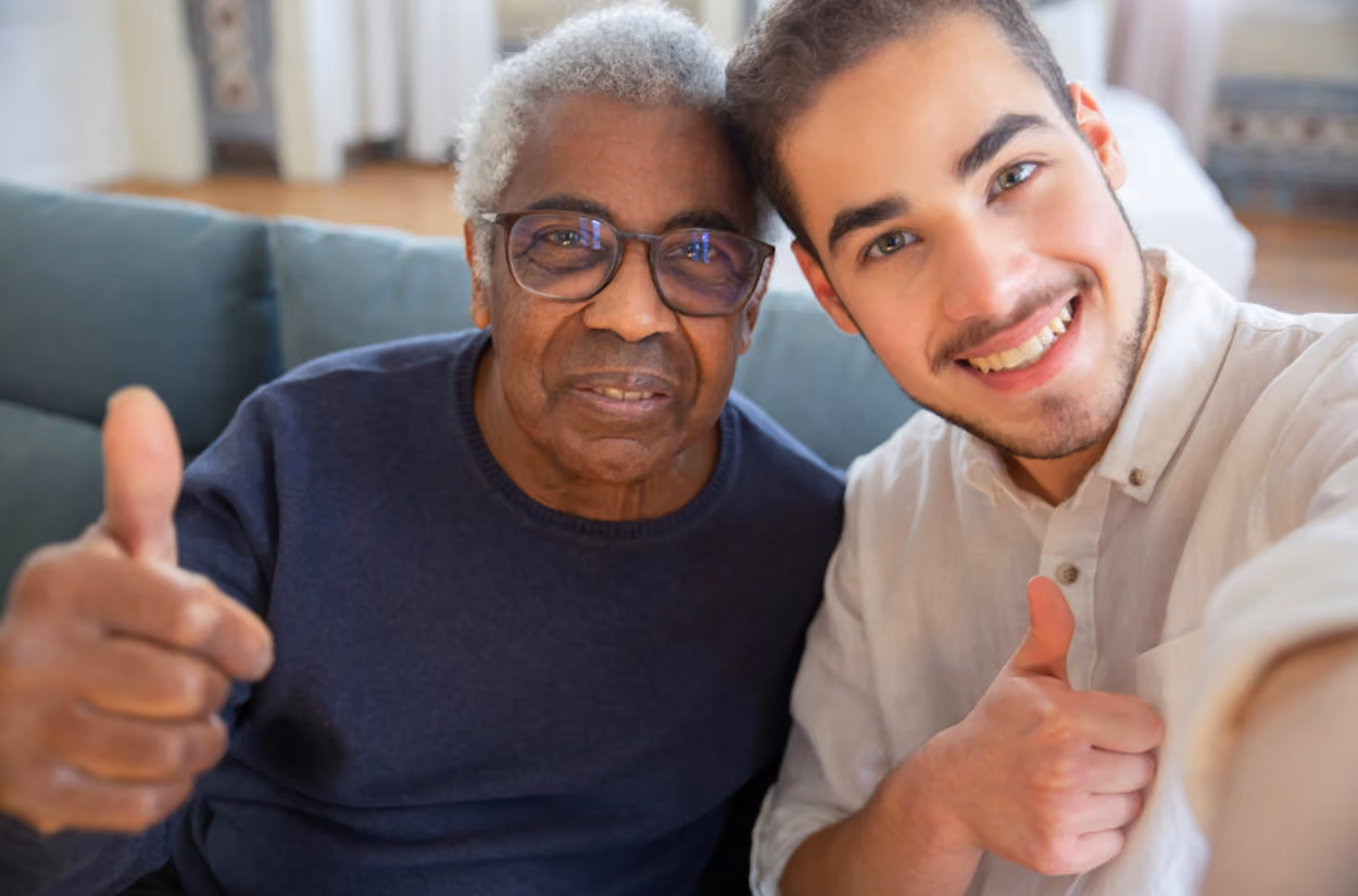 A man and an older man sit on a couch, both smiling and giving a thumbs up to the camera.