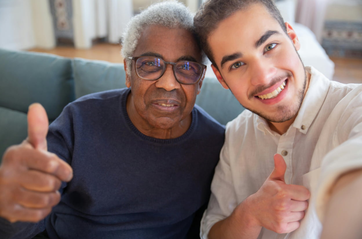 A man and an older man sit on a couch, both smiling and giving a thumbs up to the camera.