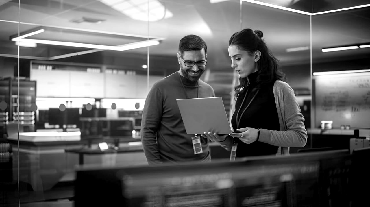 Two individuals standing together, looking at a computer screen displaying information or graphics. 