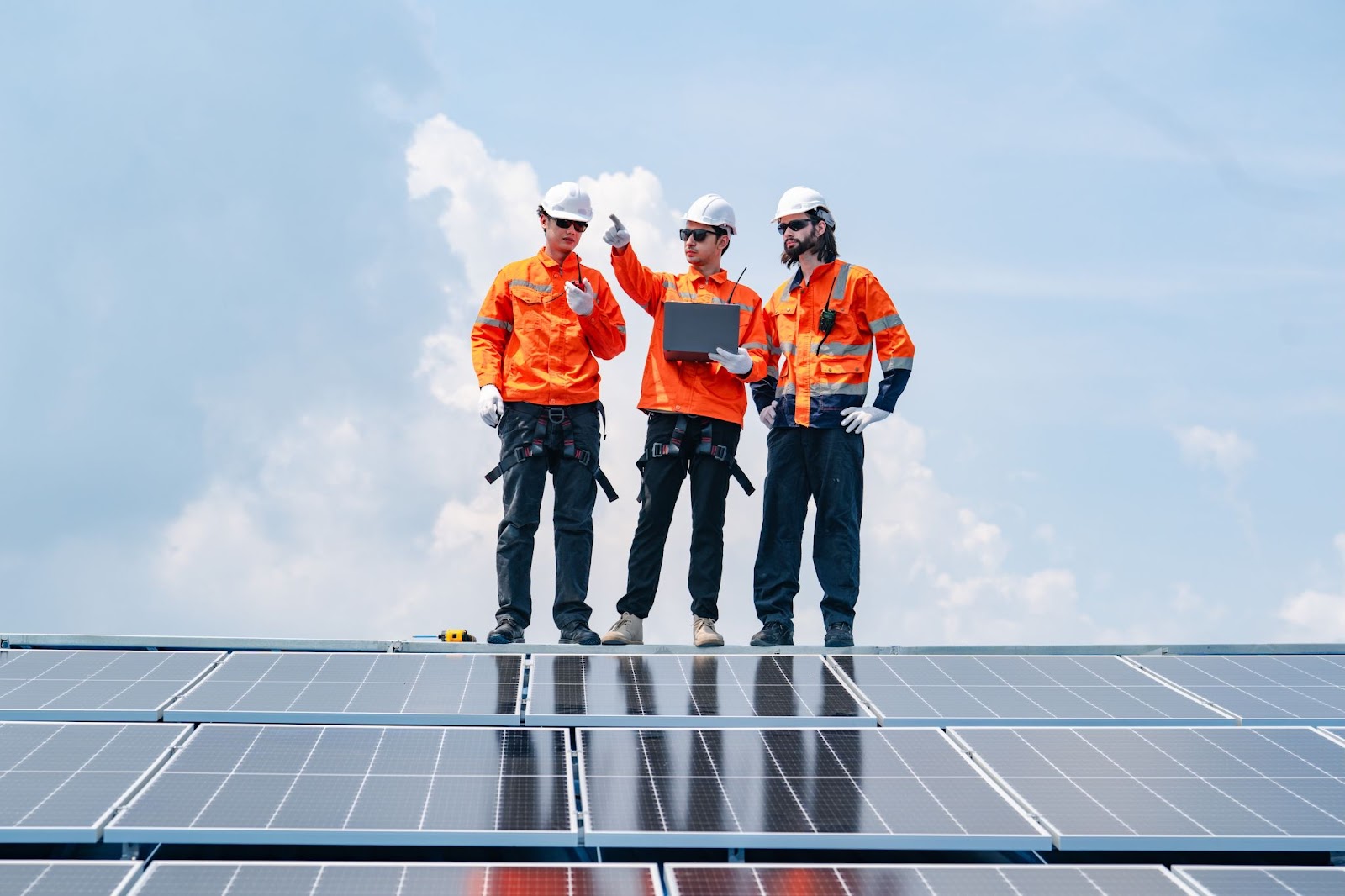 Three men in orange work clothes stand on solar panels, inspecting the installation under a clear blue sky.  