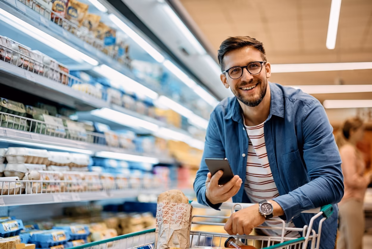 A man shopping in a grocery store holds a cell phone, possibly checking a shopping list or prices.