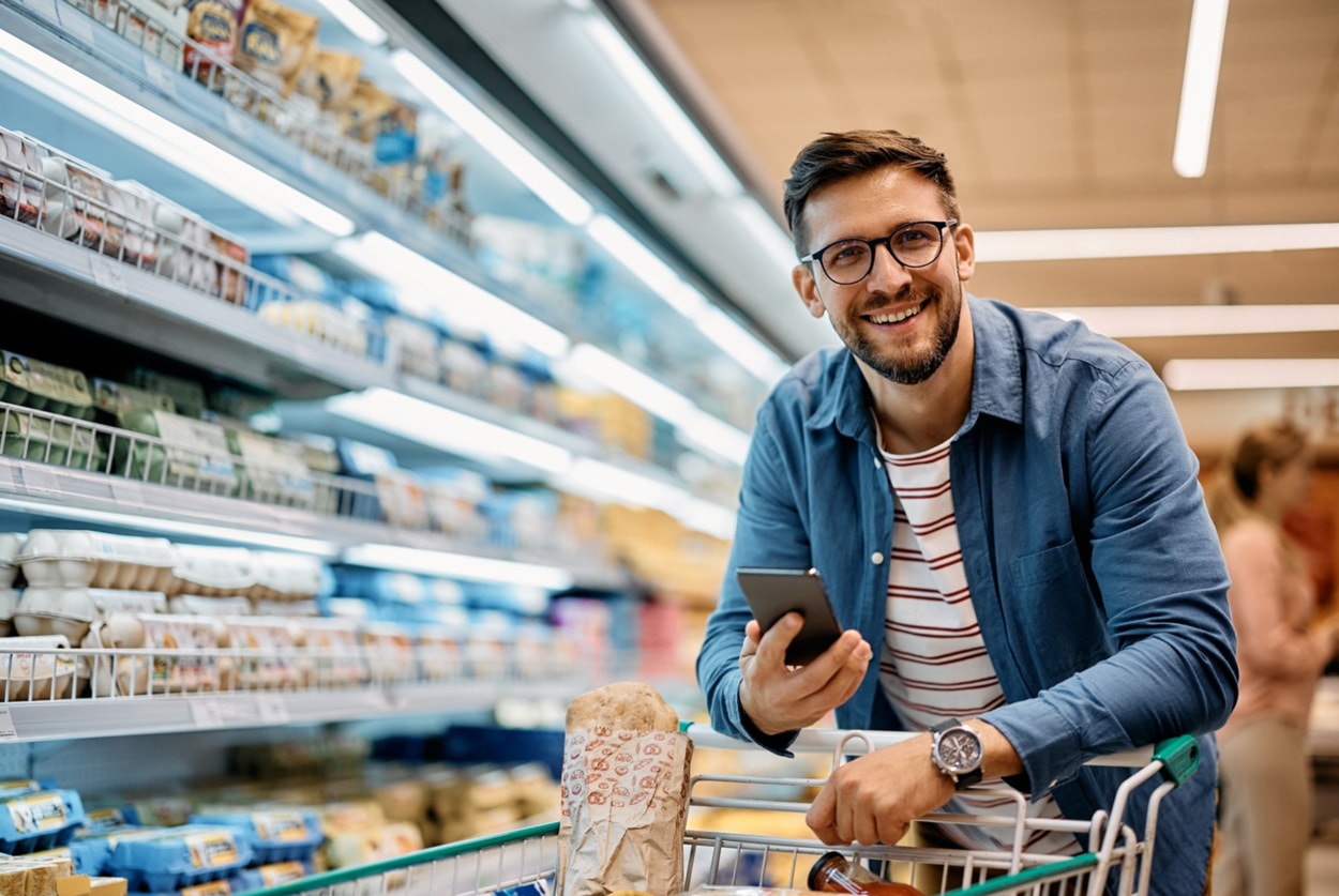 A man shopping in a grocery store holds a cell phone, possibly checking a shopping list or prices.