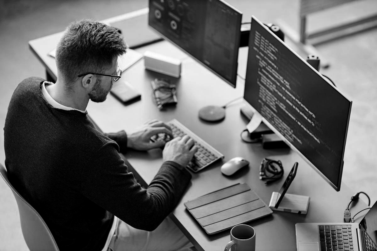 A man seated at a desk, focused on two computer monitors displaying various applications. 