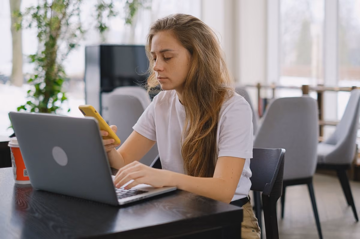 A woman seated at a table, using a laptop while a cell phone rests beside her. 