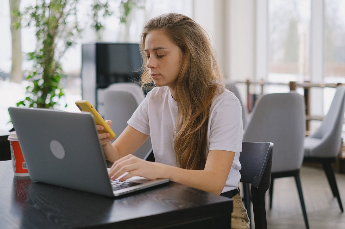 A woman seated at a table, using a laptop while a cell phone rests beside her.