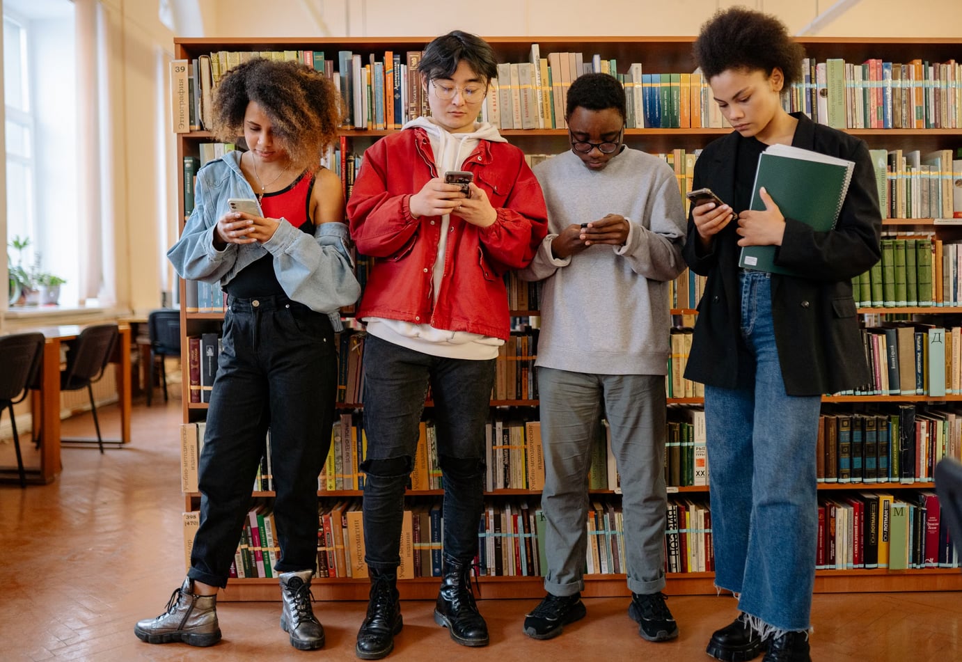 Four young individuals stand in front of bookshelves, each engaged with their smartphones, showcasing a modern library setting.  