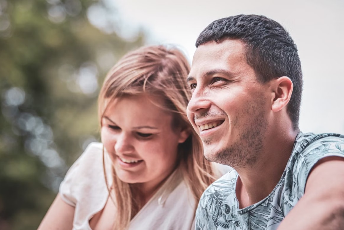 A smiling man and woman outdoors, enjoying a moment together, with blurred greenery in the background, conveying happiness.