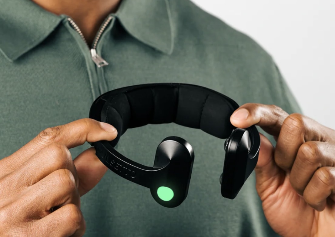 Person holding a black headband device with a glowing green light, wearing a green shirt with a zipper, against a neutral background.