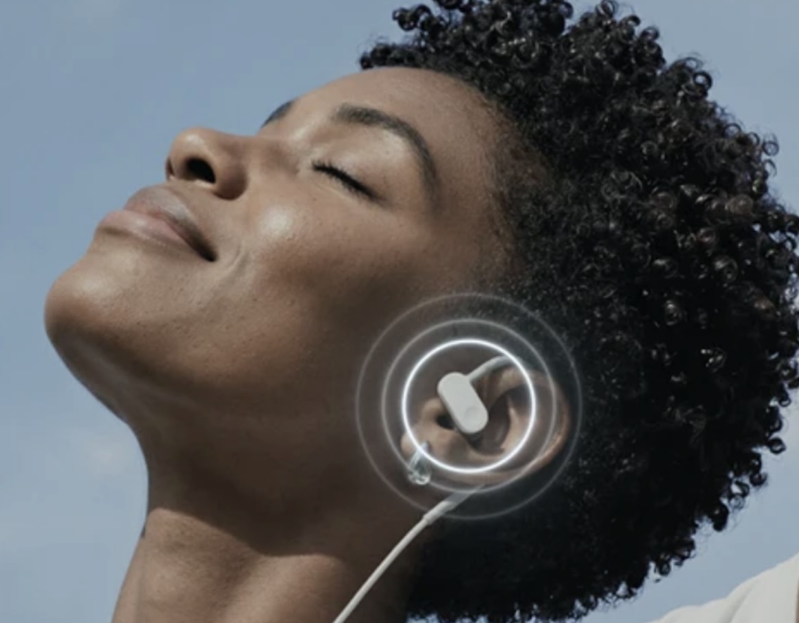 Woman with closed eyes enjoys music through wireless earbuds, highlighted by sound waves, under a clear blue sky.