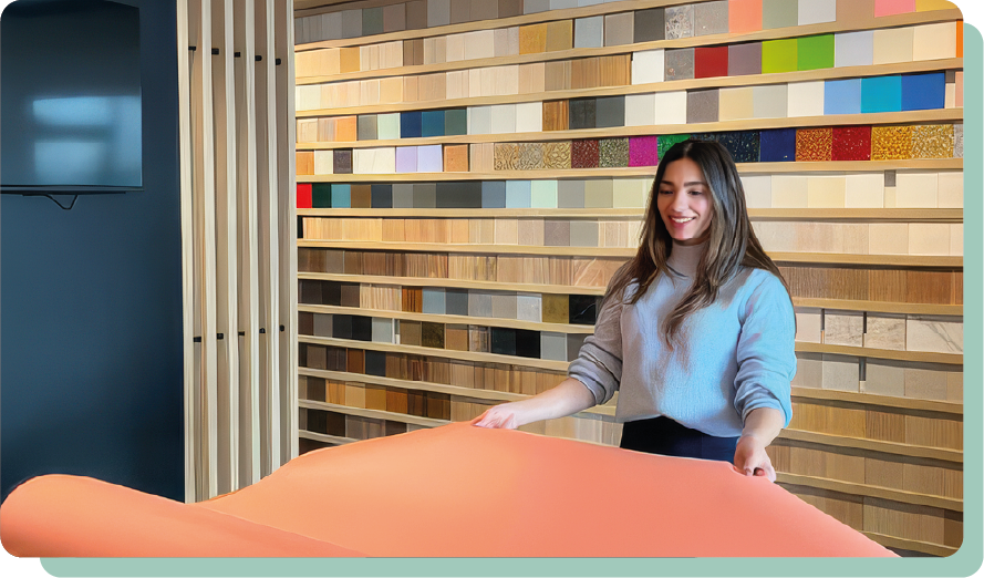 Femme souriante dans un showroom regardant un grand échantillon de tissu orange, avec un mur d'échantillons de matériaux colorés en arrière-plan.