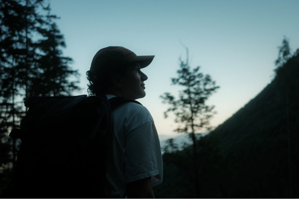 Person wearing a cap and backpack looking up in a forested mountain area at dusk.