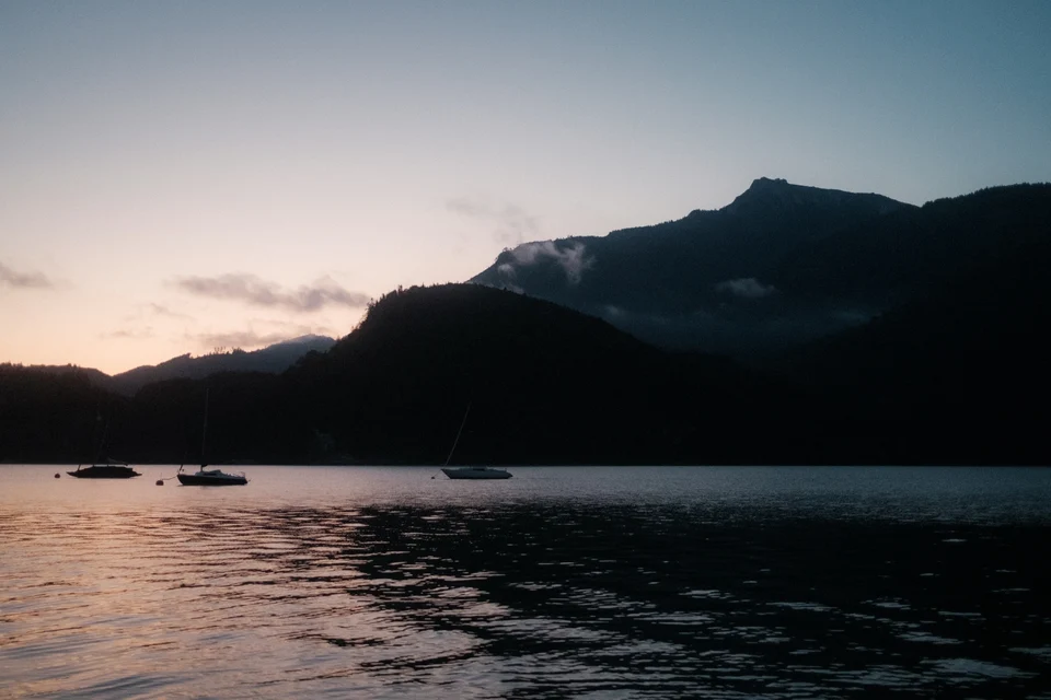 Calm lake at dusk with three sailboats anchored and silhouetted mountains in the background.
