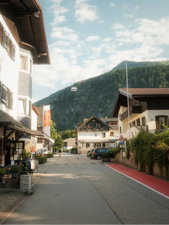 Quiet street in a small village with traditional alpine houses, parked cars, and forested mountains in the background under a partly cloudy sky.