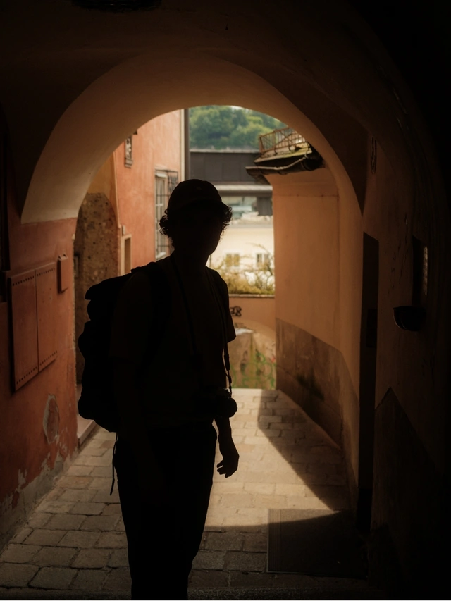 Silhouette of a person wearing a backpack and camera standing under an archway in a narrow alley.