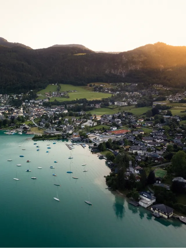 Aerial view of a lakeside town with boats anchored on calm turquoise water, surrounded by green fields and forested hills at sunset.