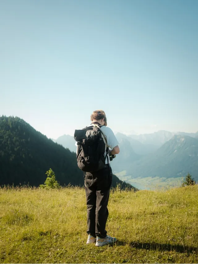 Person with a backpack standing on grassy hill overlooking a mountain valley under a clear blue sky.
