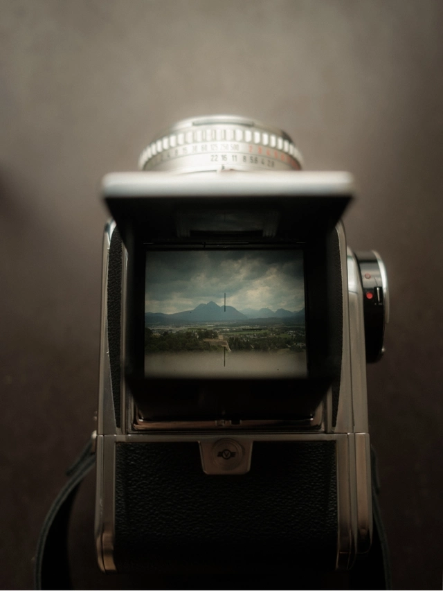 View through a vintage camera's waist-level viewfinder showing a landscape with mountains, cloudy sky, and greenery.