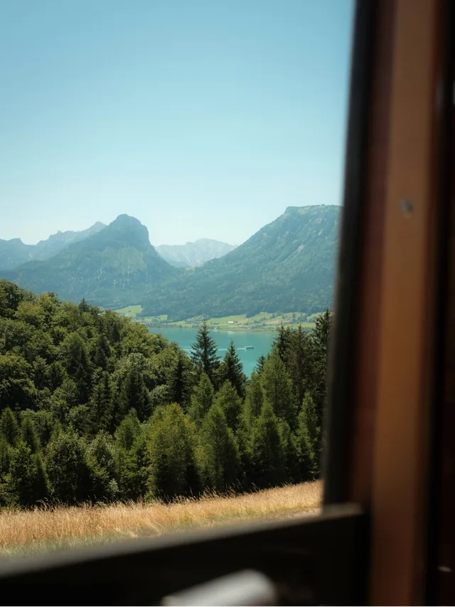View of green forested hills, a lake, and distant mountains seen through a partially open window.