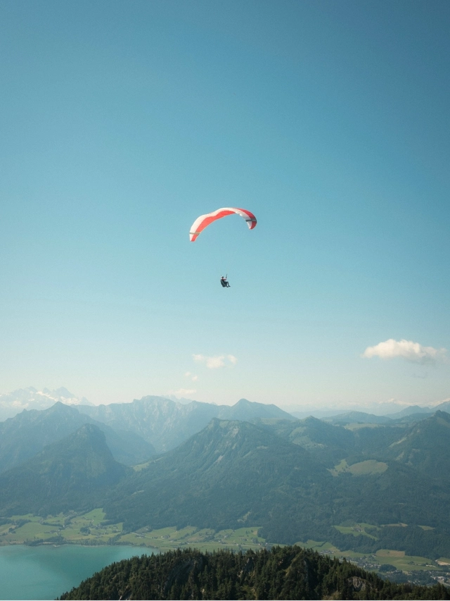 Person paragliding with a red and white parachute over mountains and a lake under a clear blue sky.