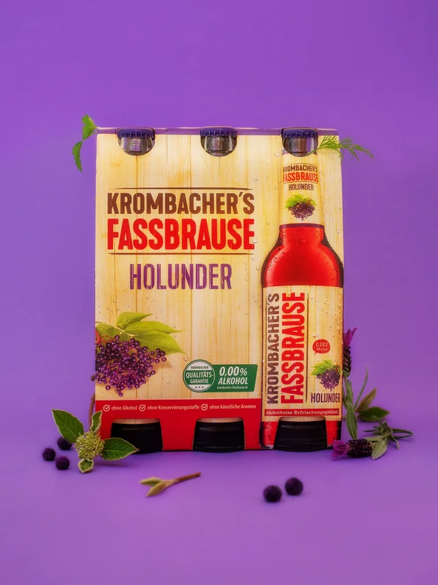 Six-pack of Krombacher's Fassbrause Holunder elderberry flavored soft drink bottles on a purple background with elderberry decoration.