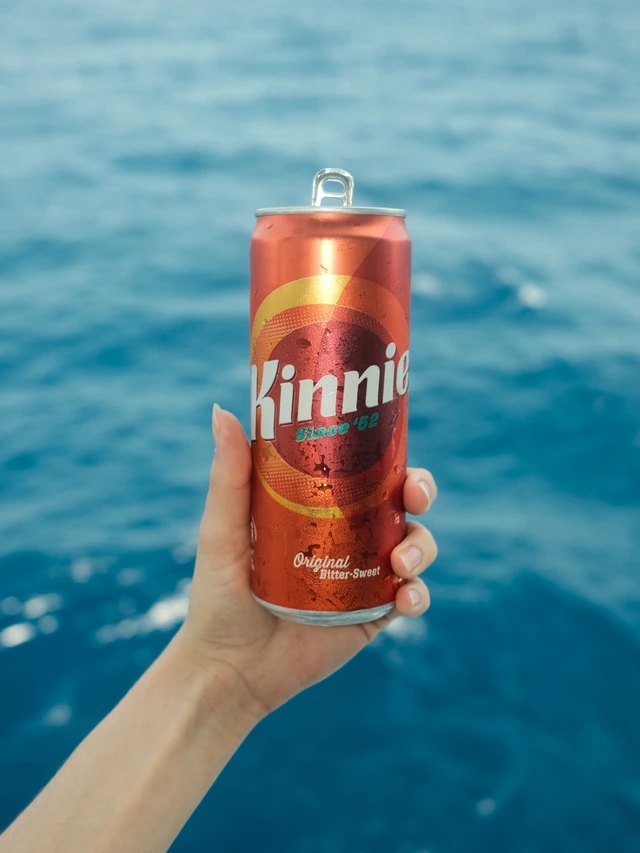 Hand holding a cold can of Kinnie soda with ocean water in the background.