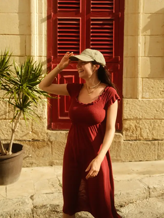 Woman in a maroon dress and green cap smiling and standing near a red shuttered window with a potted plant beside her.