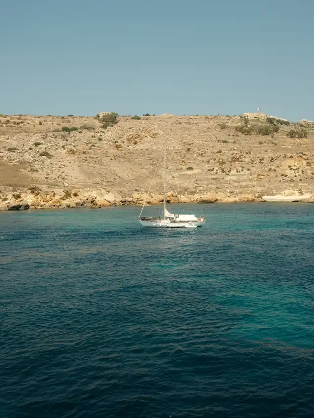 White sailboat anchored on calm blue water near a rocky, dry hillside coast under a clear sky.