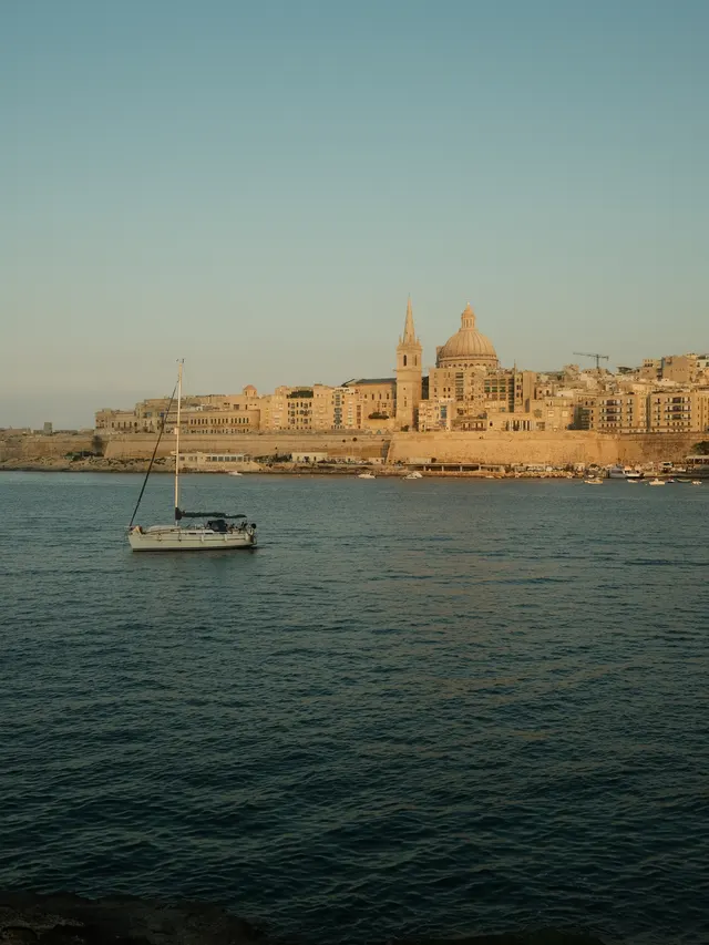 Sailboat floating on calm water with historic buildings and a large dome in the background at sunset.