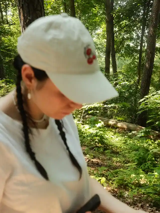 Person with braided hair wearing a white cap and t-shirt in a green forest.