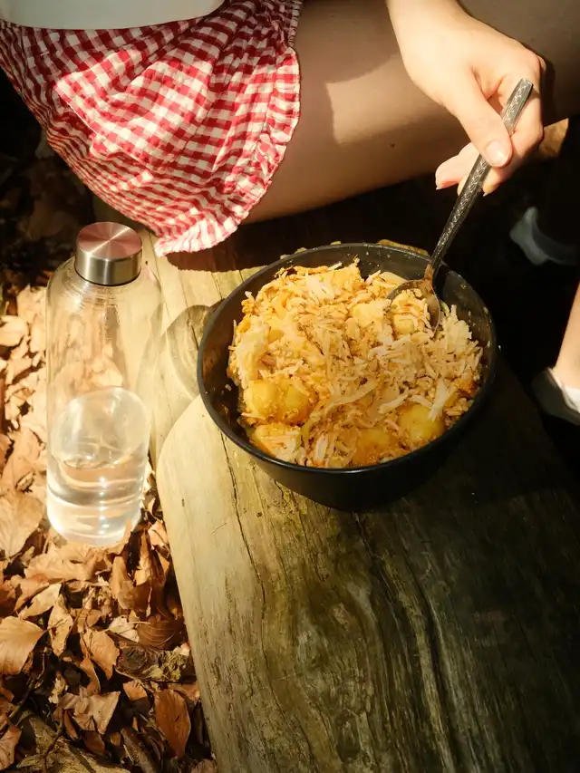 Person in red and white checkered shorts eating from a bowl of food while sitting on a wooden surface next to a clear water bottle outdoors.