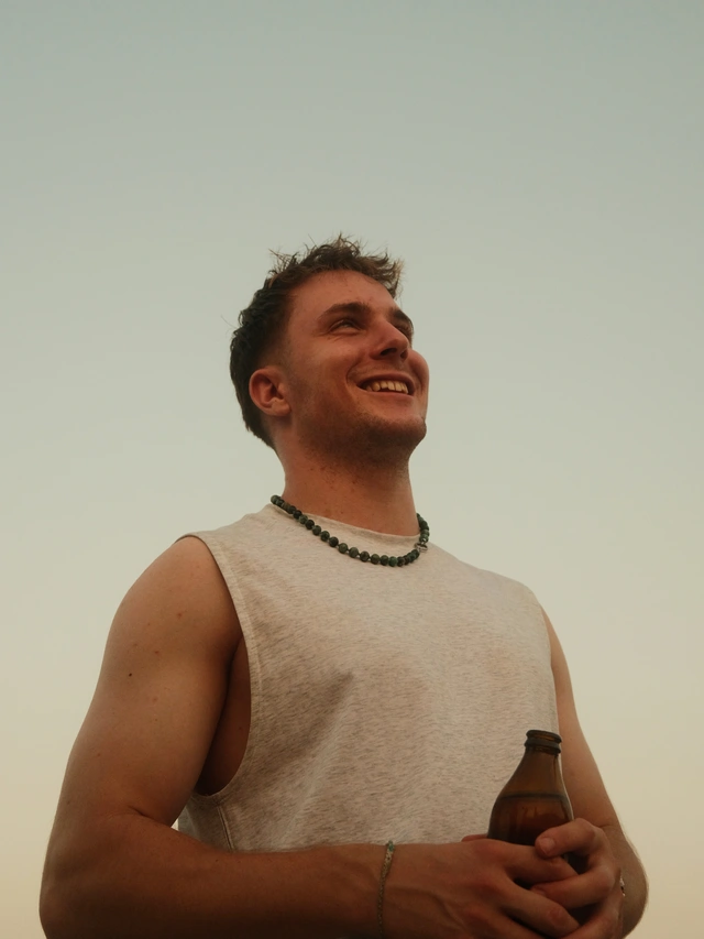 Smiling man wearing a sleeveless light gray shirt and a beaded necklace, holding a brown bottle against a clear sky.