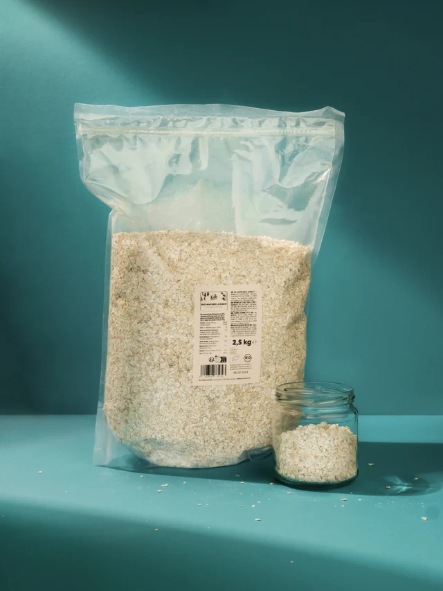 Large transparent resealable bag filled with oat flakes next to a small glass jar containing oat flakes, both on a blue surface with a blue background.