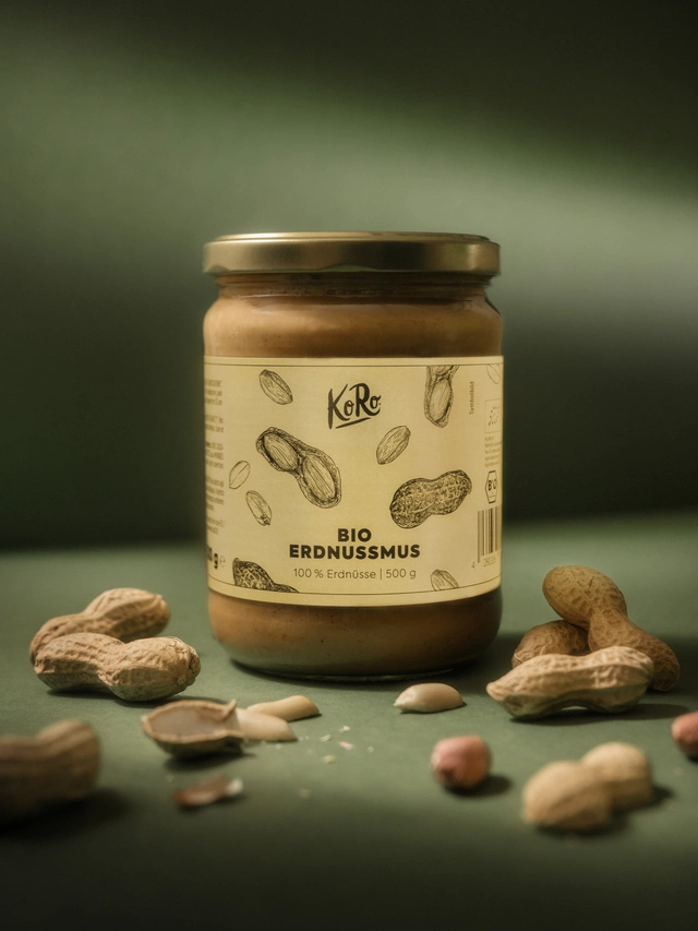 Jar of KoRo organic peanut butter surrounded by whole and shelled peanuts on a green surface.