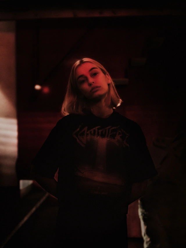 Young woman with blonde hair standing in dim red lighting wearing a dark graphic t-shirt.