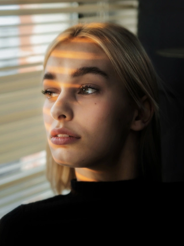 Young woman with blonde hair gazing out a window with sunlight casting striped shadows on her face.