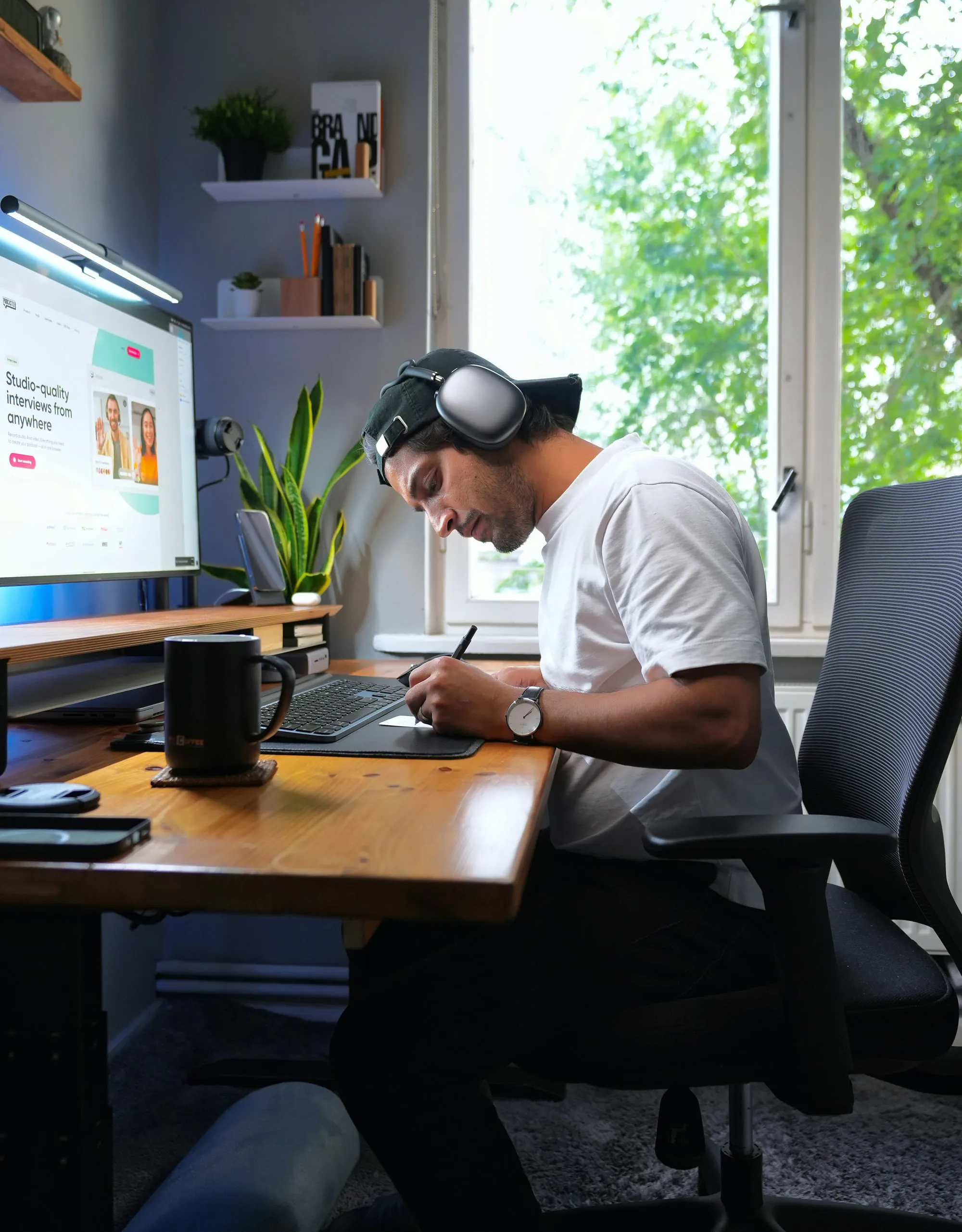 Un homme portant un casque audio et une casquette à l'envers, assis à un bureau en train d'écrire sur une tablette graphique avec un stylet, un grand écran d'ordinateur devant lui affiche une interface d'entretien de qualité studio.