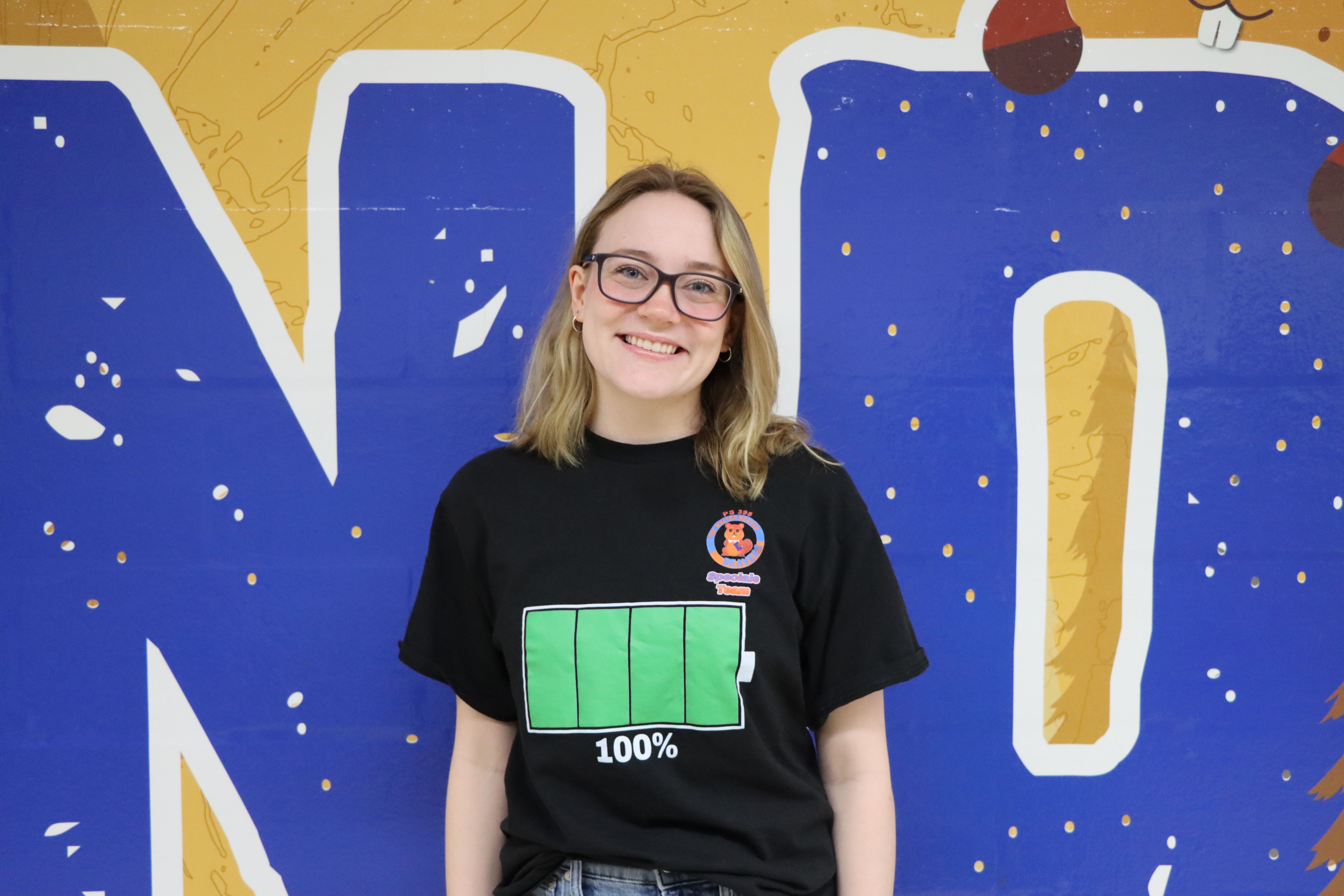 Student wearing black shirt with battery graphic smiling in front of blue mural