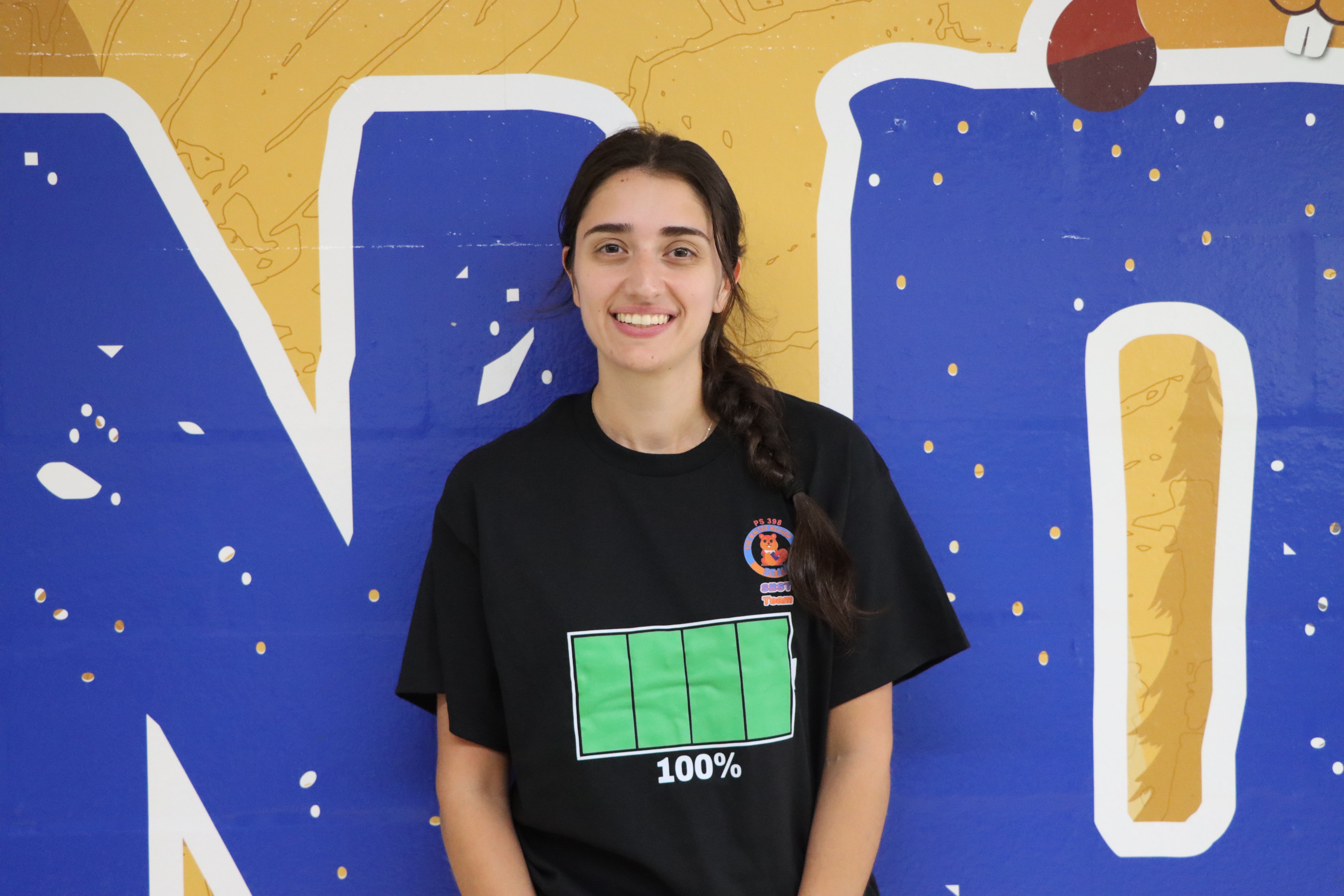 Student wearing battery graphic shirt smiles in front of blue mural